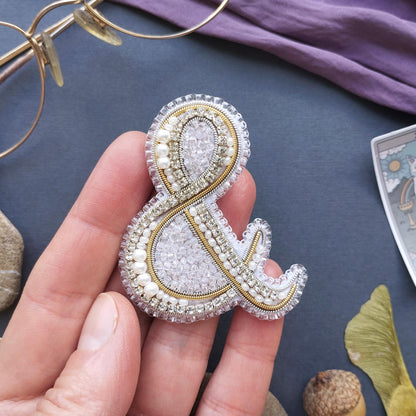 A hand holding a decorative beaded letter '&' on a dark background, with various other items such as glasses, a card, and a leaf visible in the background.
