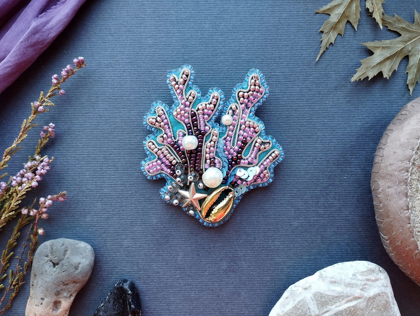 a decorative piece of jewelry, possibly a brooch or pin, adorned with various colorful beads and pearls, placed on a blue surface surrounded by natural elements such as leaves and rocks.