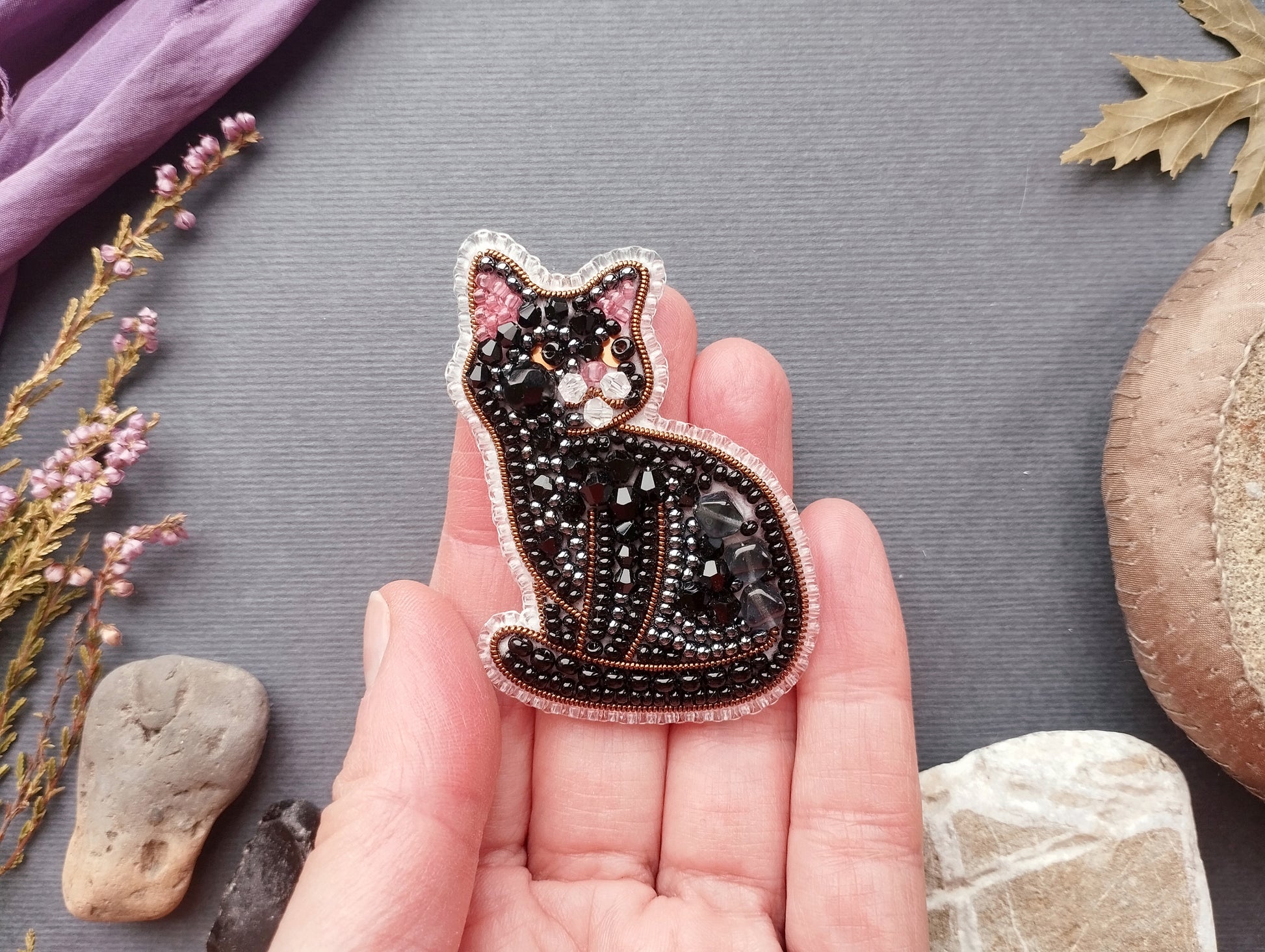 A hand holding a decorative black cat pin with beaded details against a gray background.