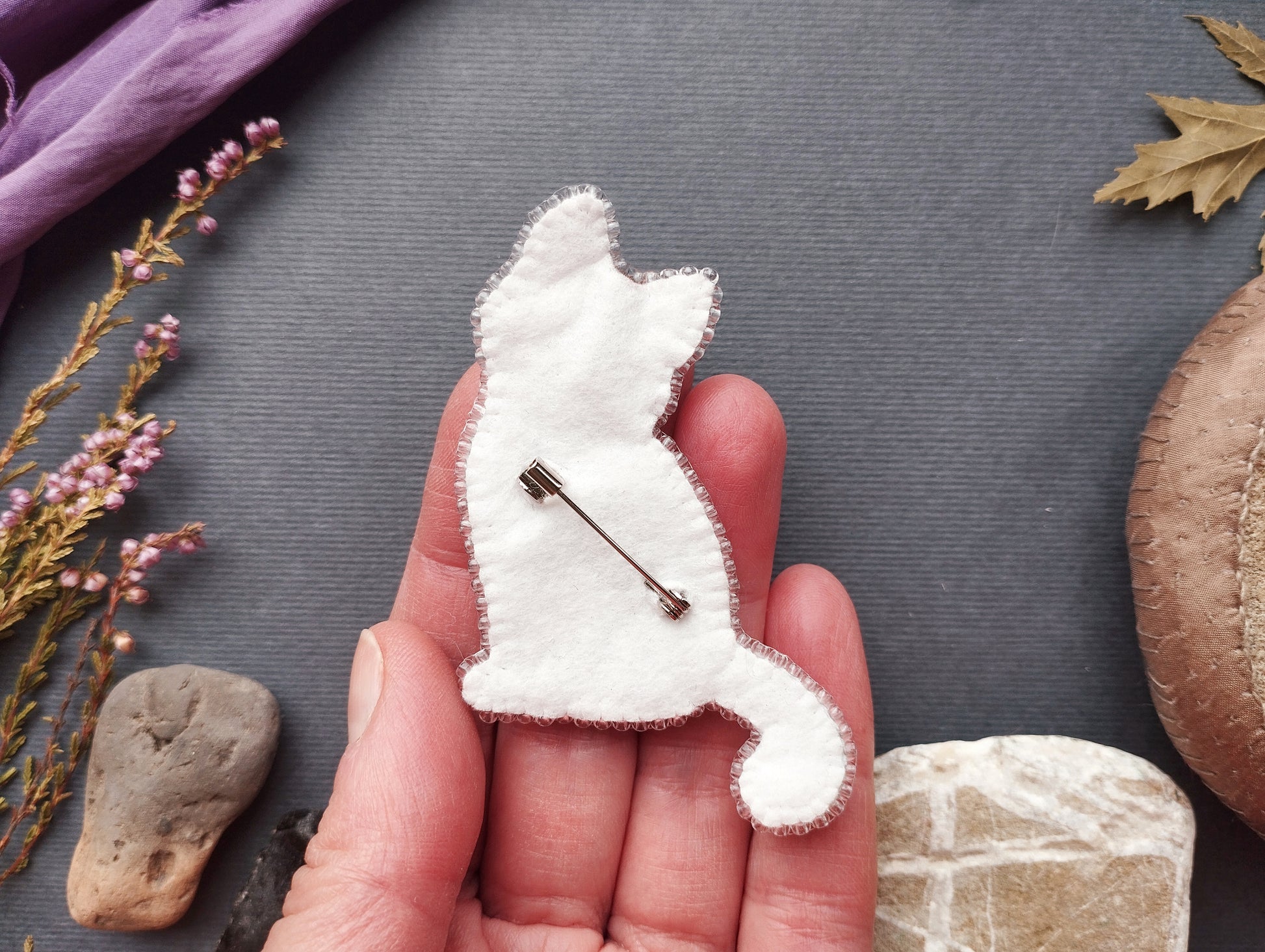A hand holding a white felt cat pin against a dark background with various natural elements, such as leaves and rocks.