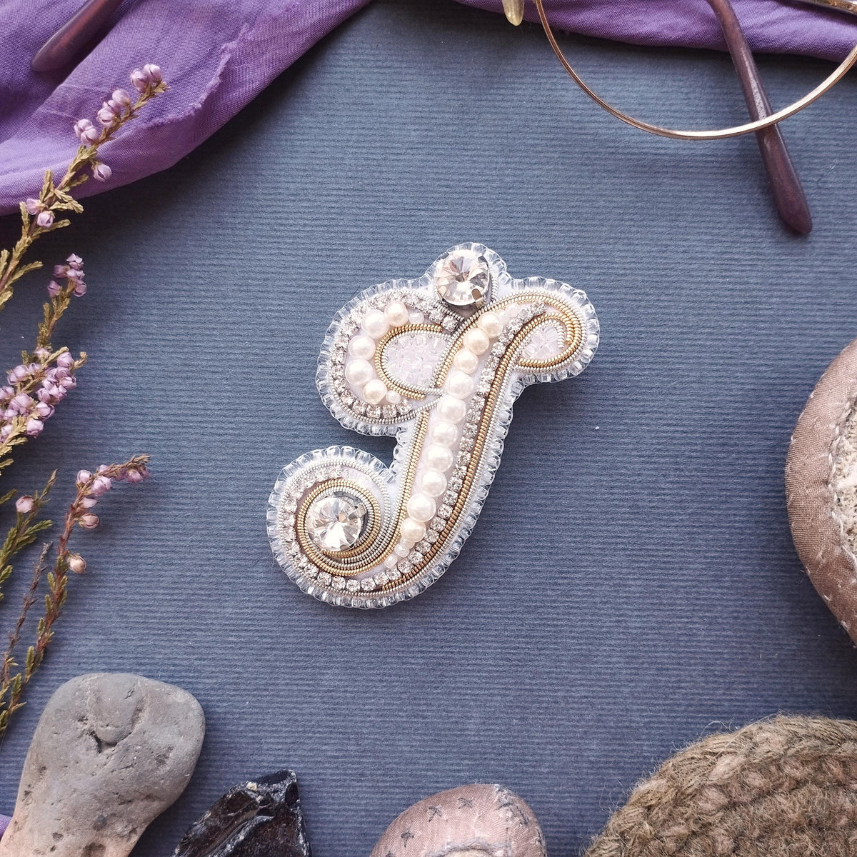 a decorative monogrammed letter 'F' adorned with pearls and rhinestones, surrounded by various natural elements such as flowers, rocks, and a pair of glasses.