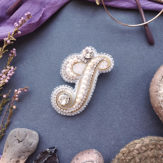 a decorative monogrammed letter 'F' adorned with pearls and rhinestones, surrounded by various natural elements such as flowers, rocks, and a pair of glasses.