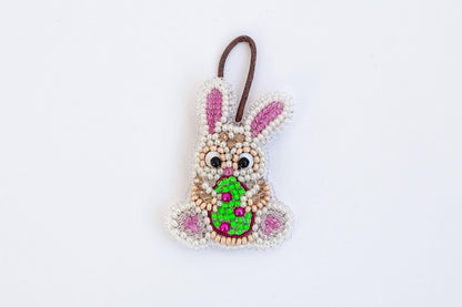 A beaded Easter bunny ornament with a green egg in its mouth, hanging against a white background.
