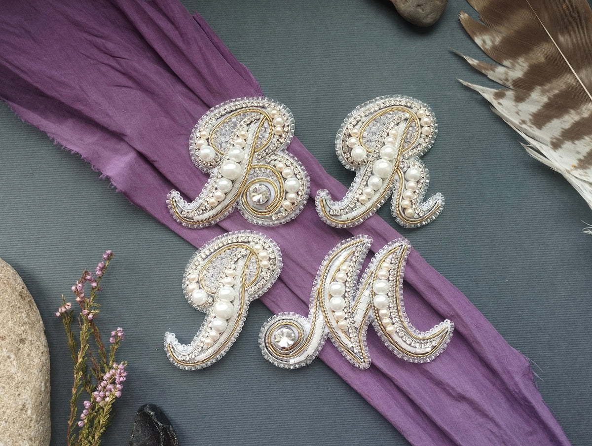 a collection of decorative elements, including a purple fabric with the letters "BR" and "PM" made of beads and pearls, a feather, and some small flowers.