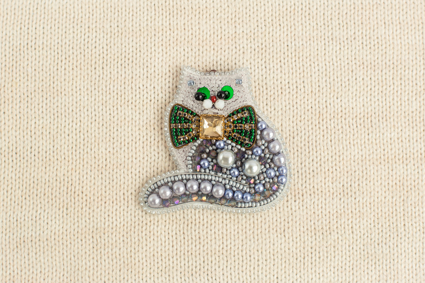 A decorative cat brooch made of various materials, including beads, pearls, and a green bow tie.