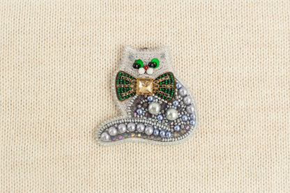 A decorative cat brooch made of various materials, including beads, pearls, and a green bow tie.