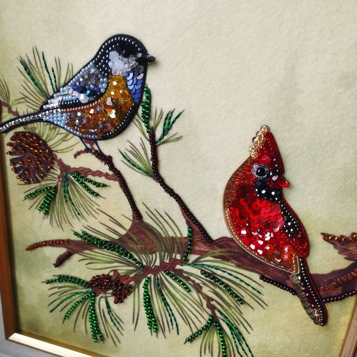 The image is a detailed embroidered piece featuring two birds perched on a branch, with pine needles and pine cones as part of the design.
