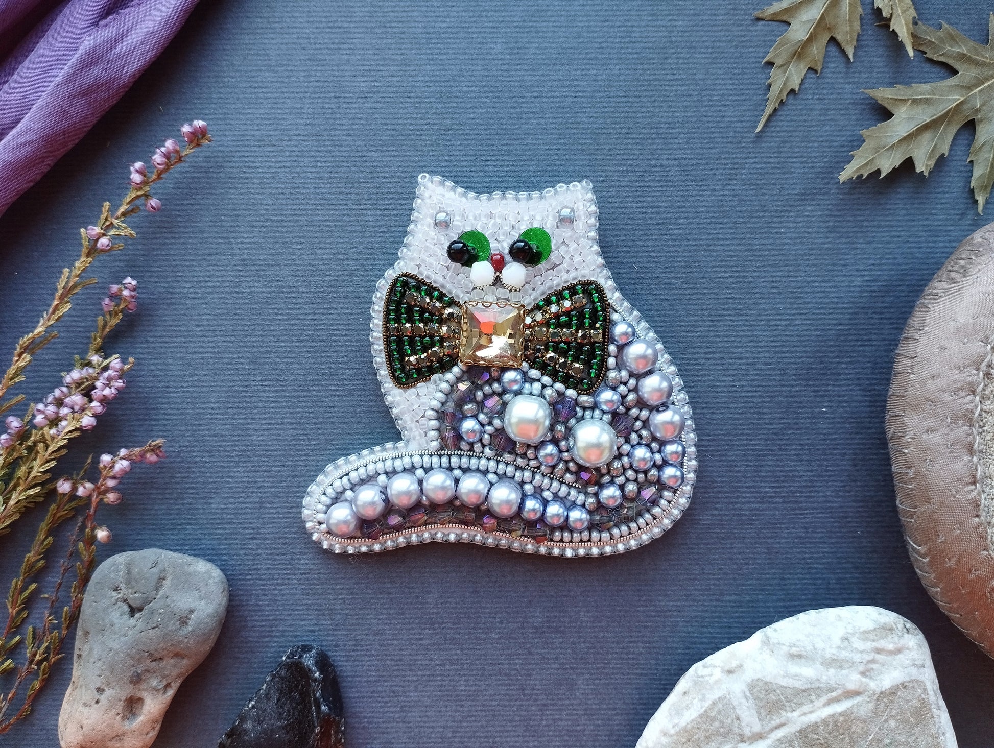 a decorative cat-shaped brooch made of beads and pearls, placed on a blue surface surrounded by various natural elements such as leaves, rocks, and flowers.