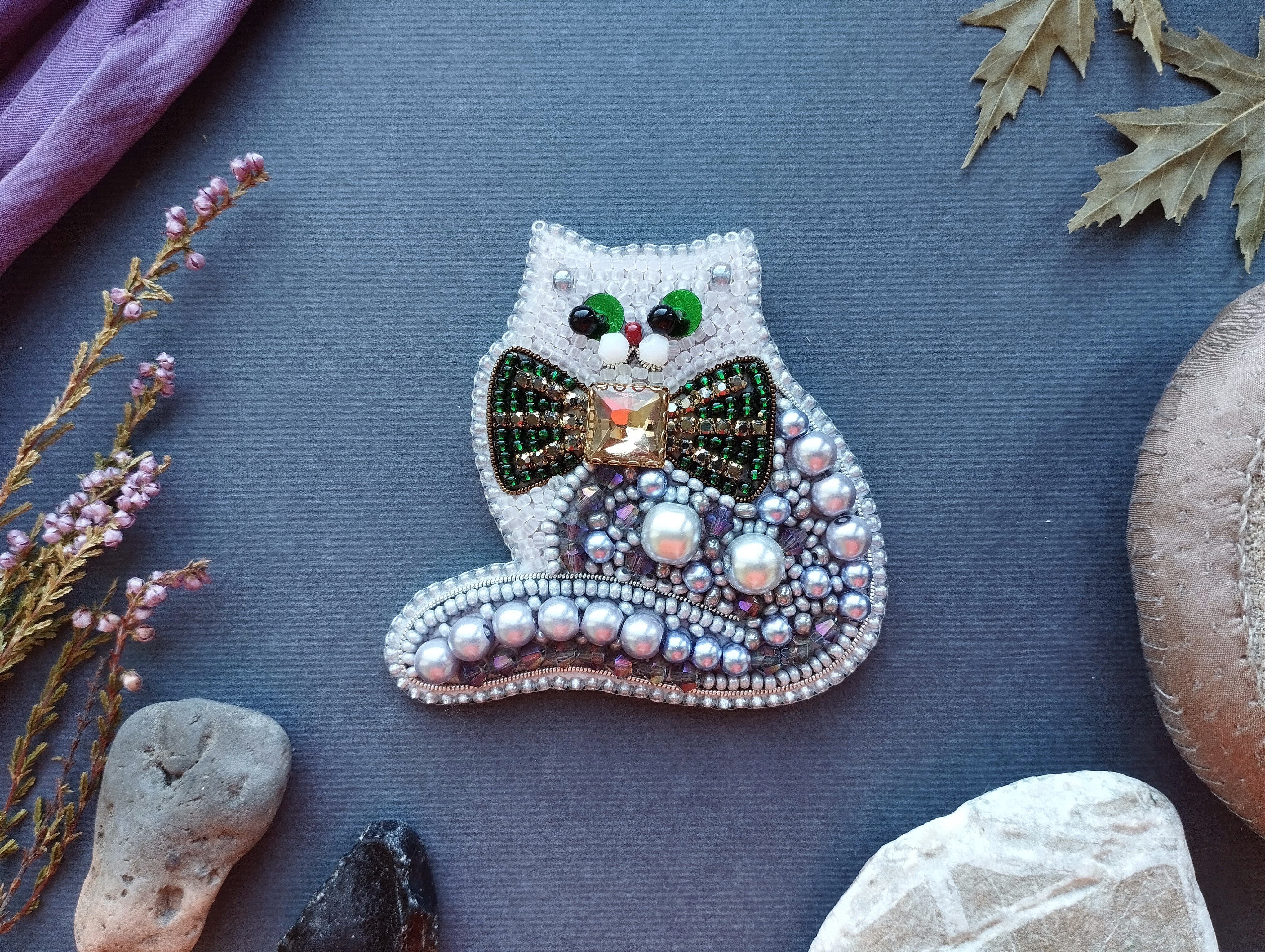a decorative cat-shaped brooch made of beads and pearls, placed on a blue surface surrounded by various natural elements such as leaves, rocks, and flowers.