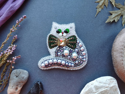 a decorative cat-shaped brooch made of beads and pearls, placed on a blue surface surrounded by various natural elements such as leaves, rocks, and flowers.