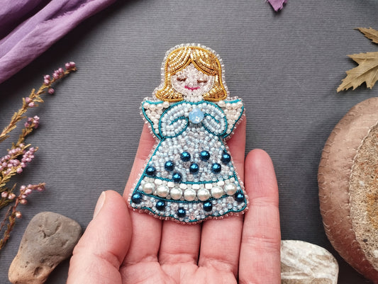A hand holding a decorative beaded doll with a blue dress and pearl accents, surrounded by various natural elements such as flowers, leaves, and rocks.