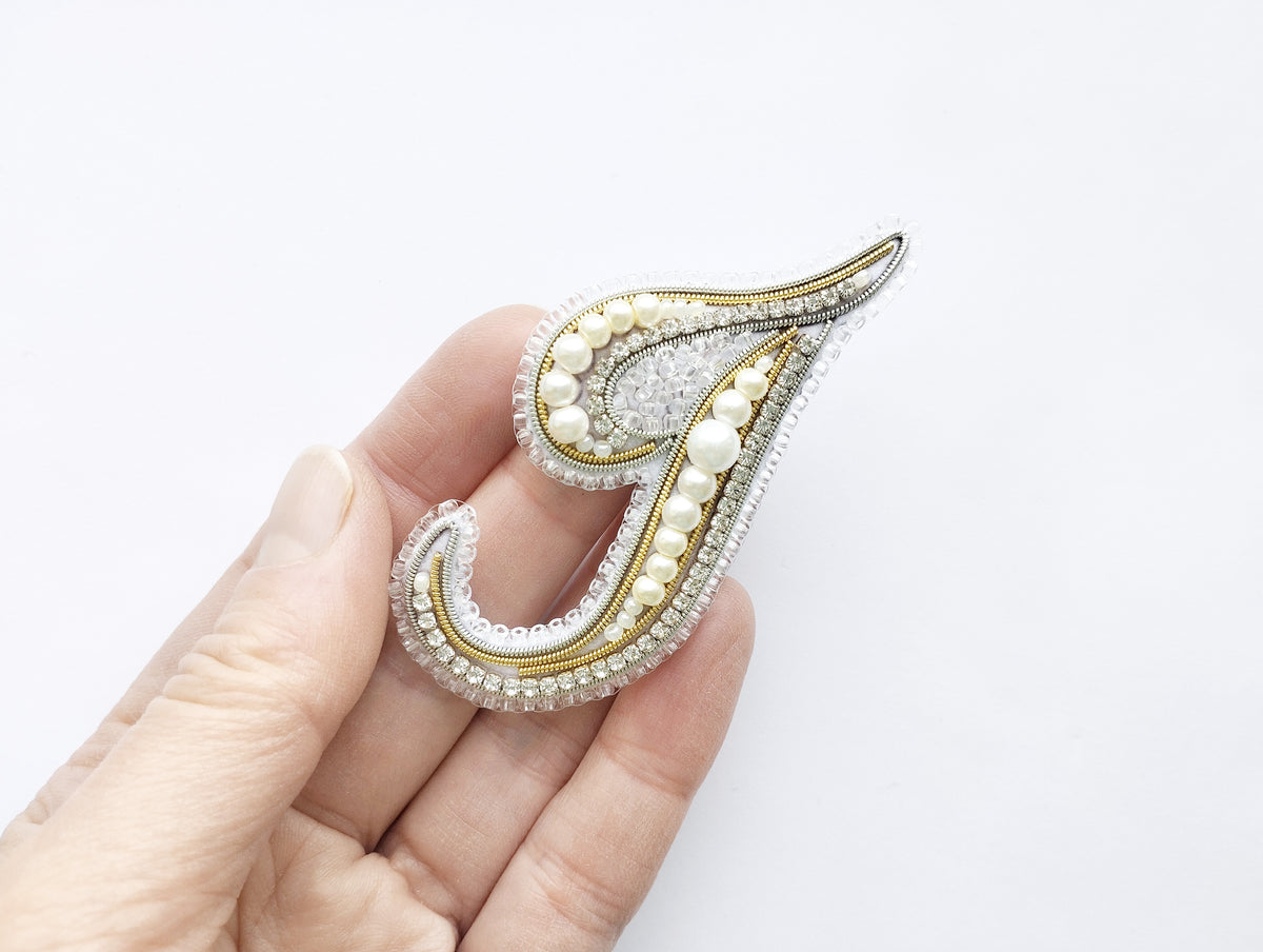 A hand holding a decorative piece of jewelry, which appears to be a brooch or pin with a floral design and pearls.