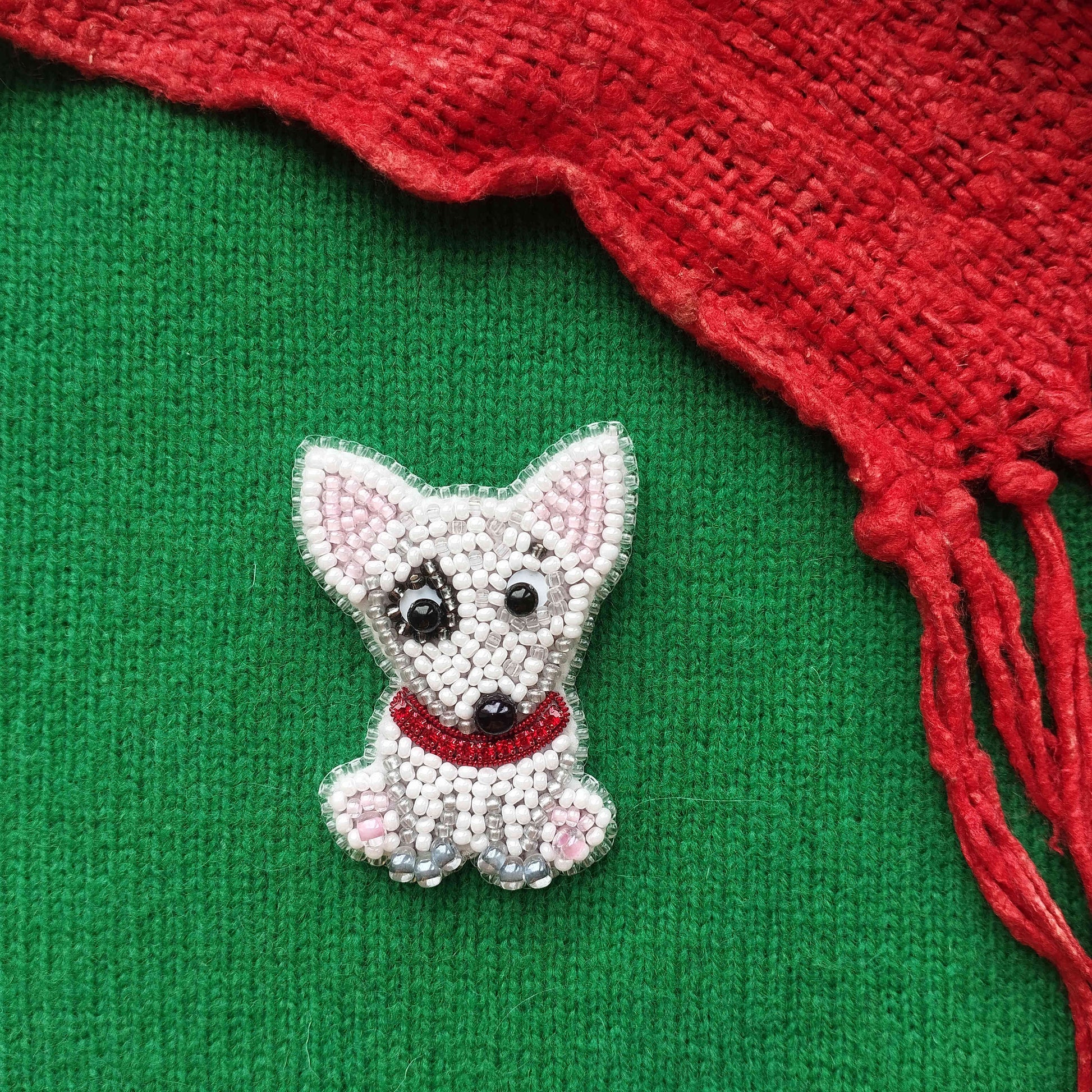 A small, white, beaded dog-shaped brooch is displayed on a green background, with a red, knitted or crocheted item partially visible to the right.