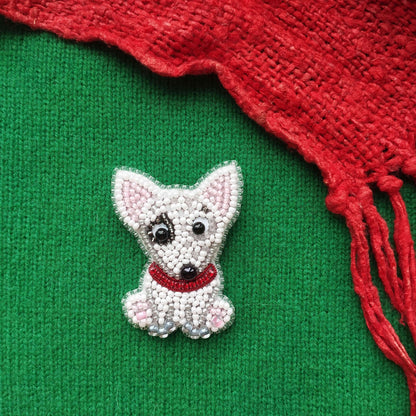 A small, white, beaded dog-shaped brooch is displayed on a green background, with a red, knitted or crocheted item partially visible to the right.