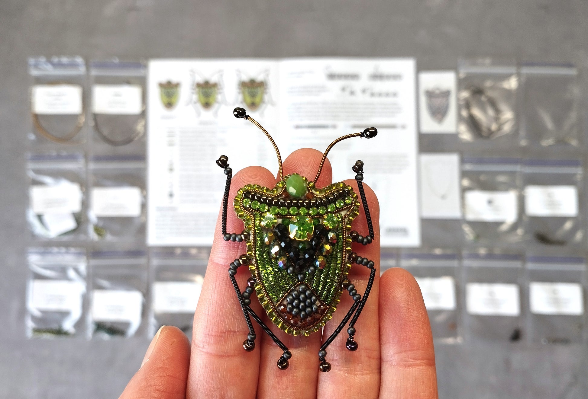 A hand is holding a green and black beaded insect brooch against a background of various bags and papers.