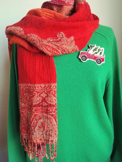 A person wearing a green sweater with a red and orange patterned scarf draped over it, and a small patch on the sweater featuring a red truck.