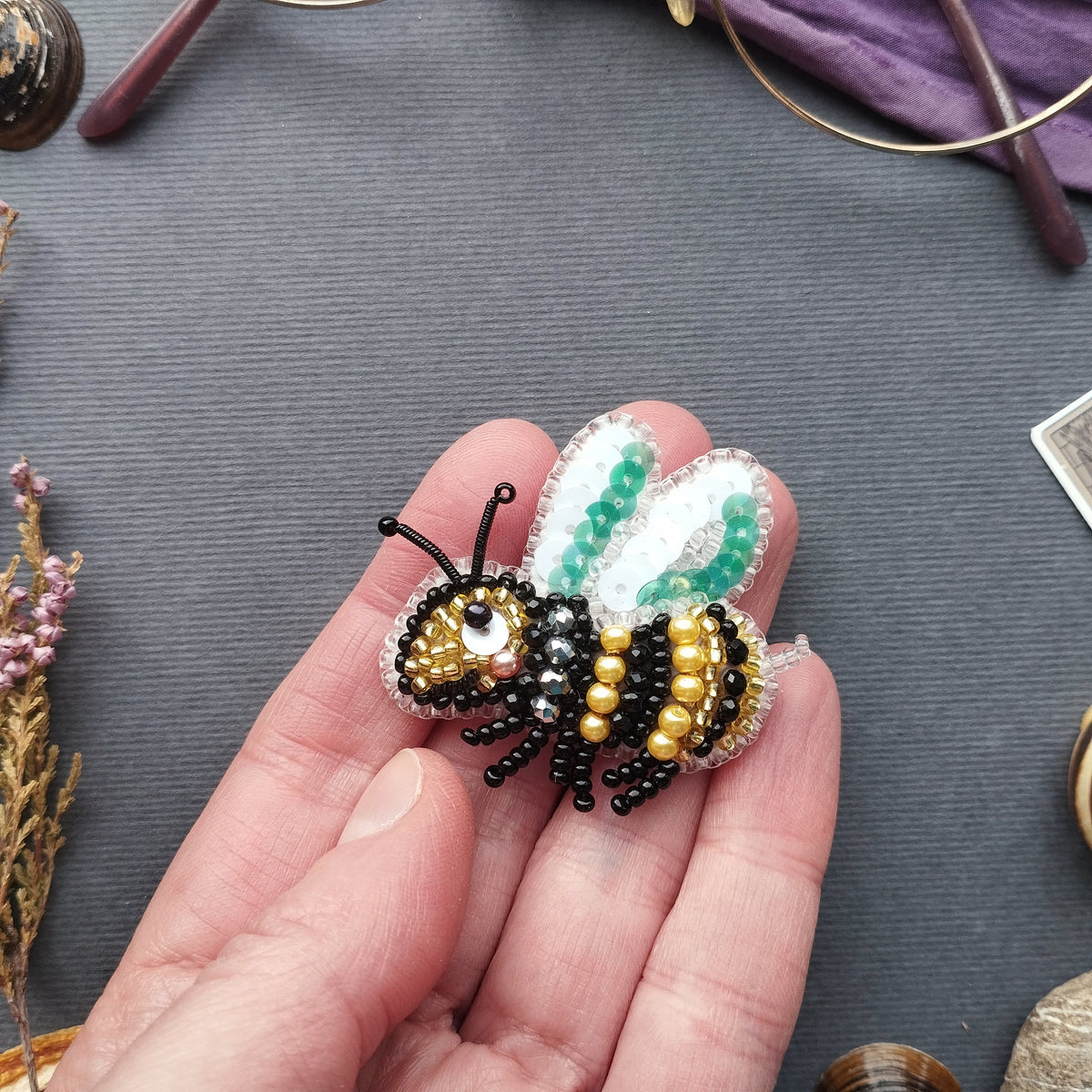 A hand holding a small, intricately beaded bee brooch against a gray background.
