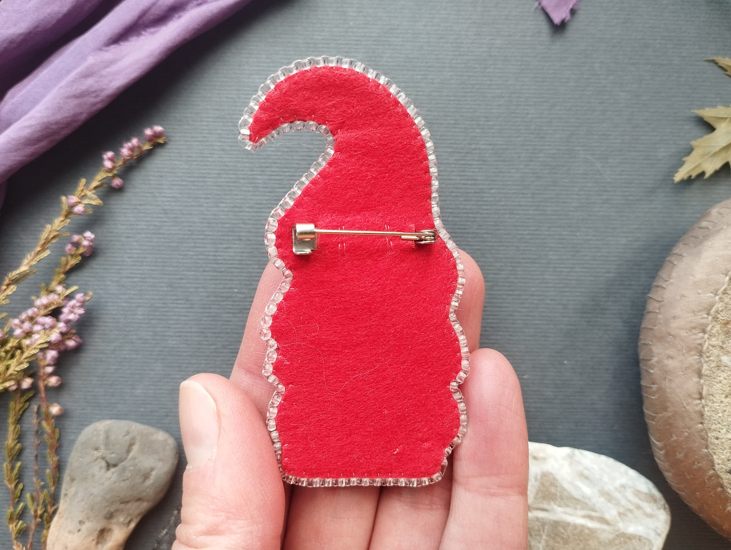 A hand holding a red felt pin or pin-like object with a silver pin attached to it, against a background of various objects such as leaves, rocks, and flowers.