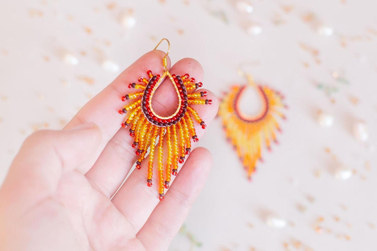 A hand holding a pair of vibrant, beaded earrings against a blurred background of more beaded earrings.
