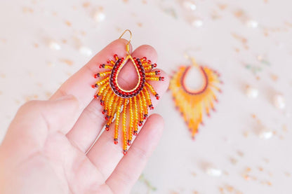 A hand holding a pair of vibrant, beaded earrings against a blurred background of more beaded earrings.