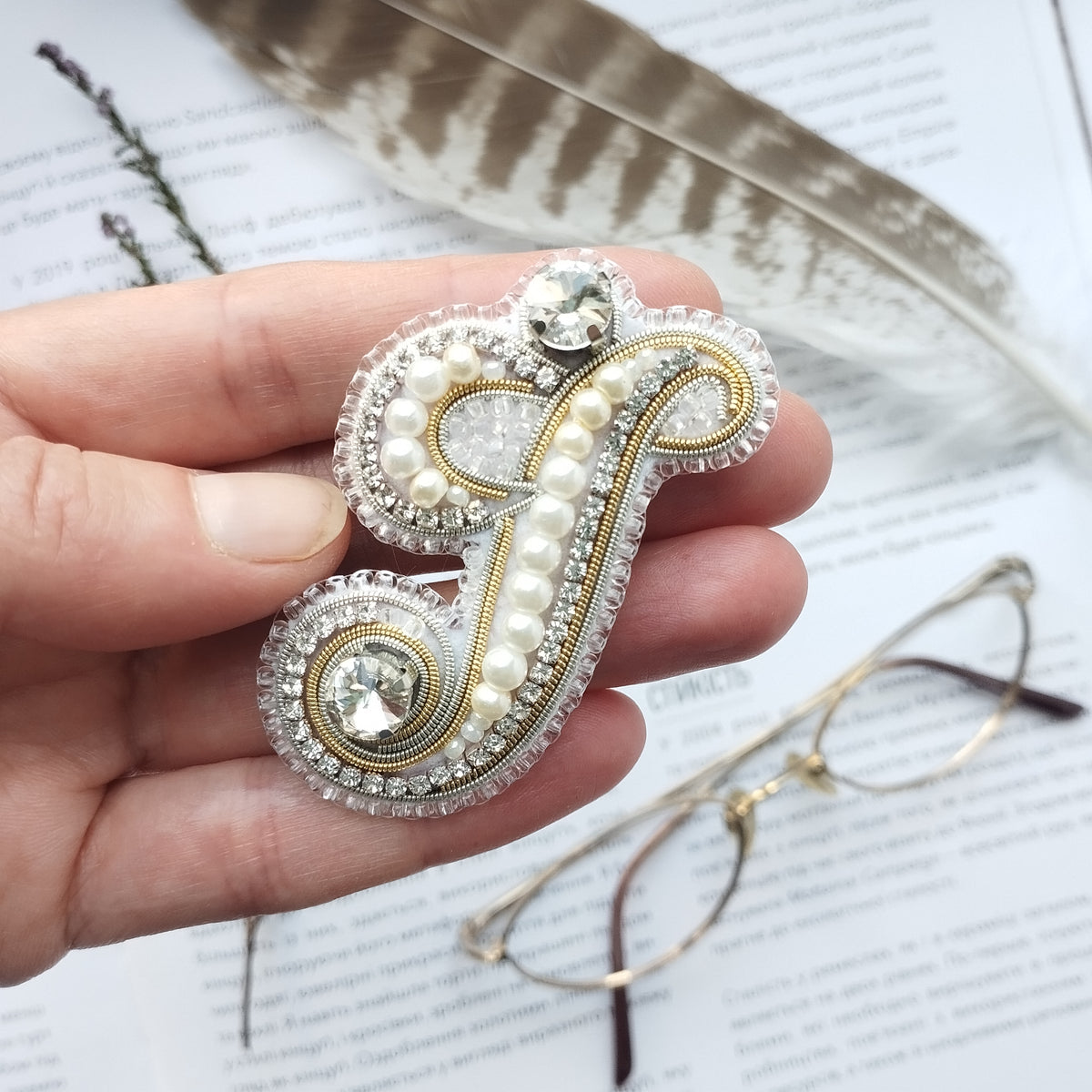 A hand holding a decorative brooch with pearls and rhinestones, with a feather and glasses in the background.