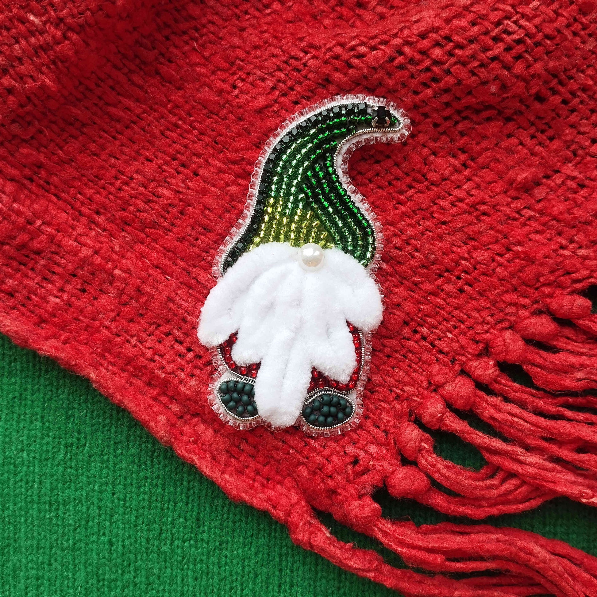 A red knitted scarf with a green and white gnome-shaped brooch made of beads and sequins is displayed on a green surface.
