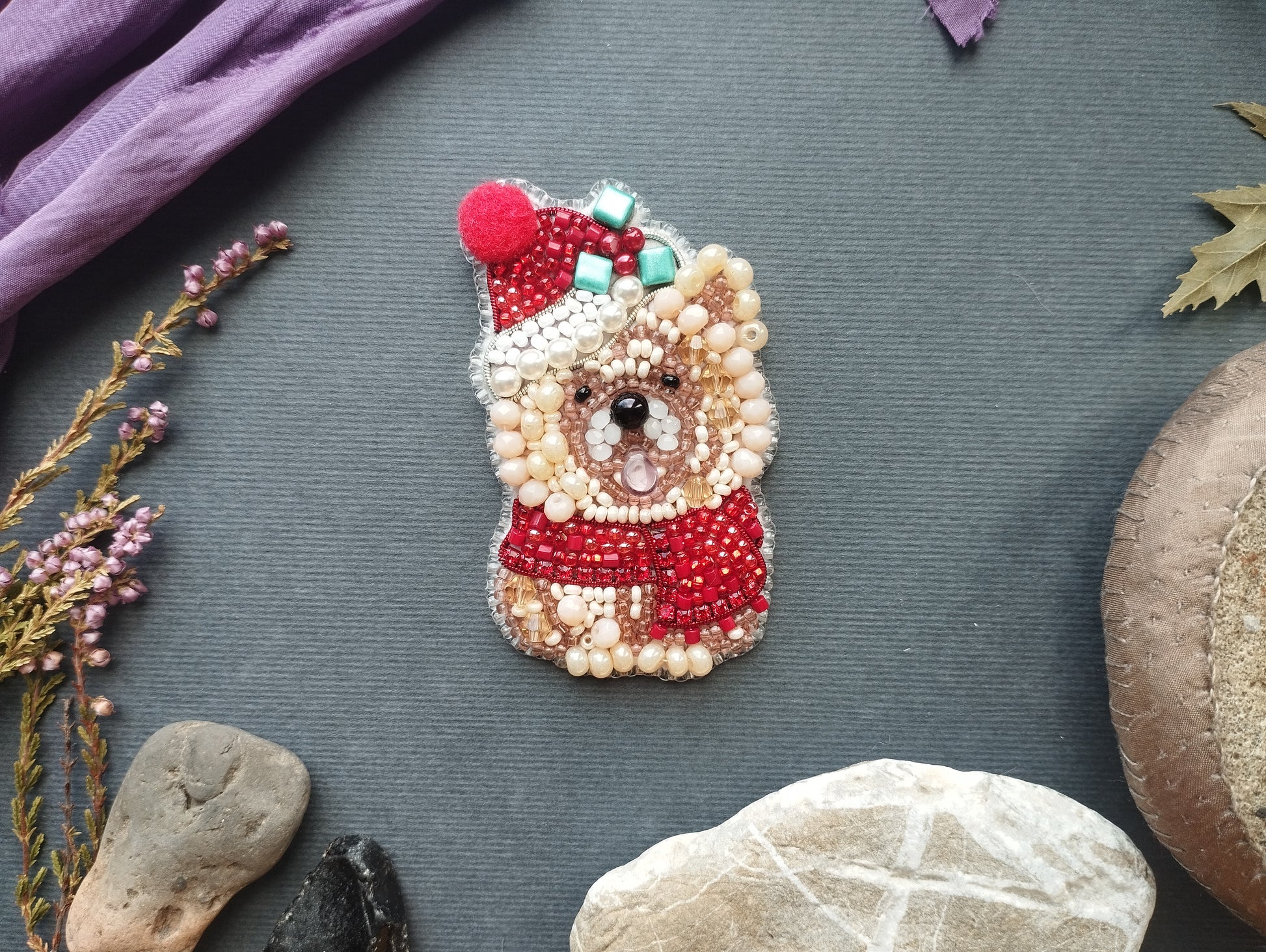 a small, intricately beaded dog pin or brooch, adorned with red and white beads, placed on a gray surface. Surrounding the dog pin are various objects, including a purple fabric, a small leaf, and some rocks.