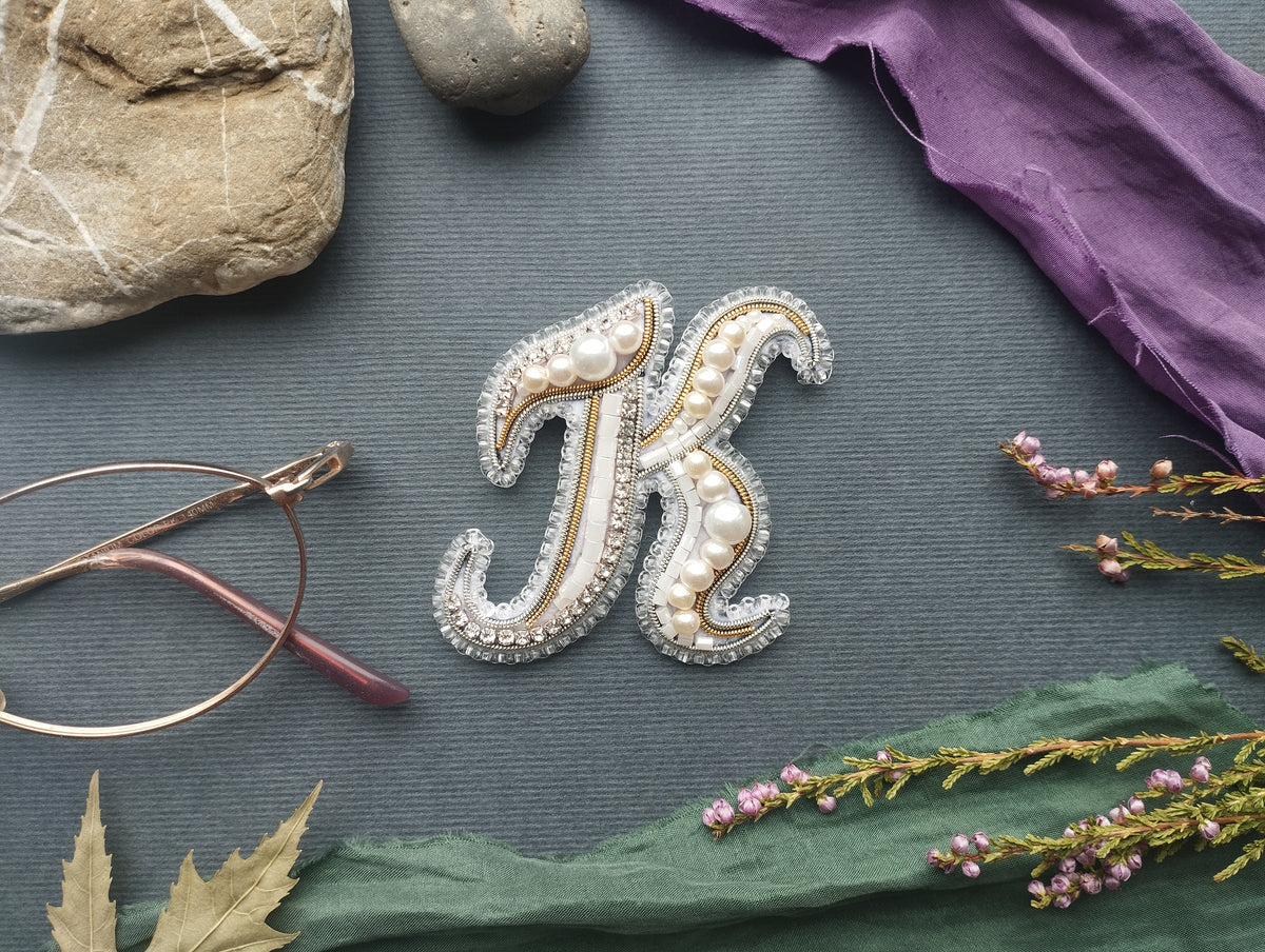 a close-up view of a decorative letter "K" made of pearls and beads, surrounded by various objects such as a rock, a pair of glasses, and some flowers.
