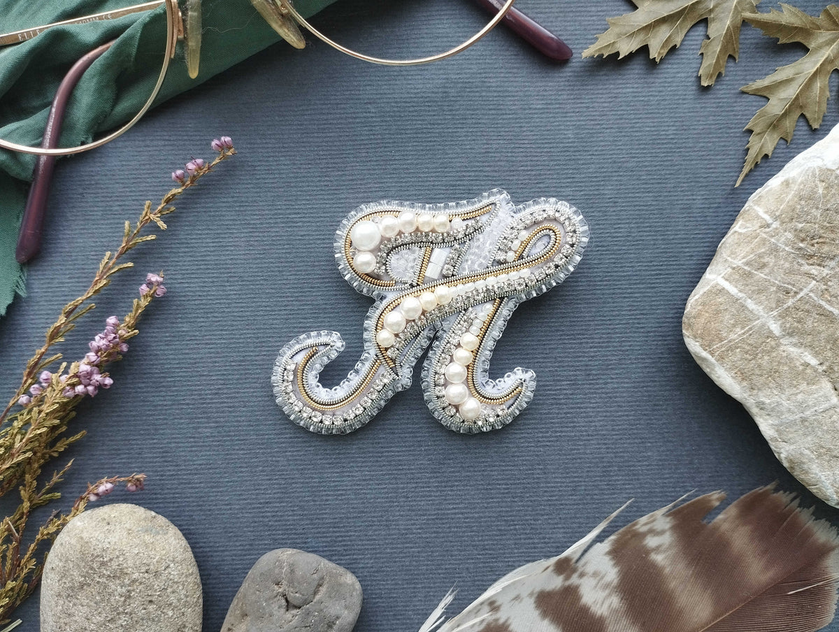 a close-up view of a decorative monogrammed letter 'N' made of silver and white beads, surrounded by various natural elements such as leaves, feathers, and rocks.