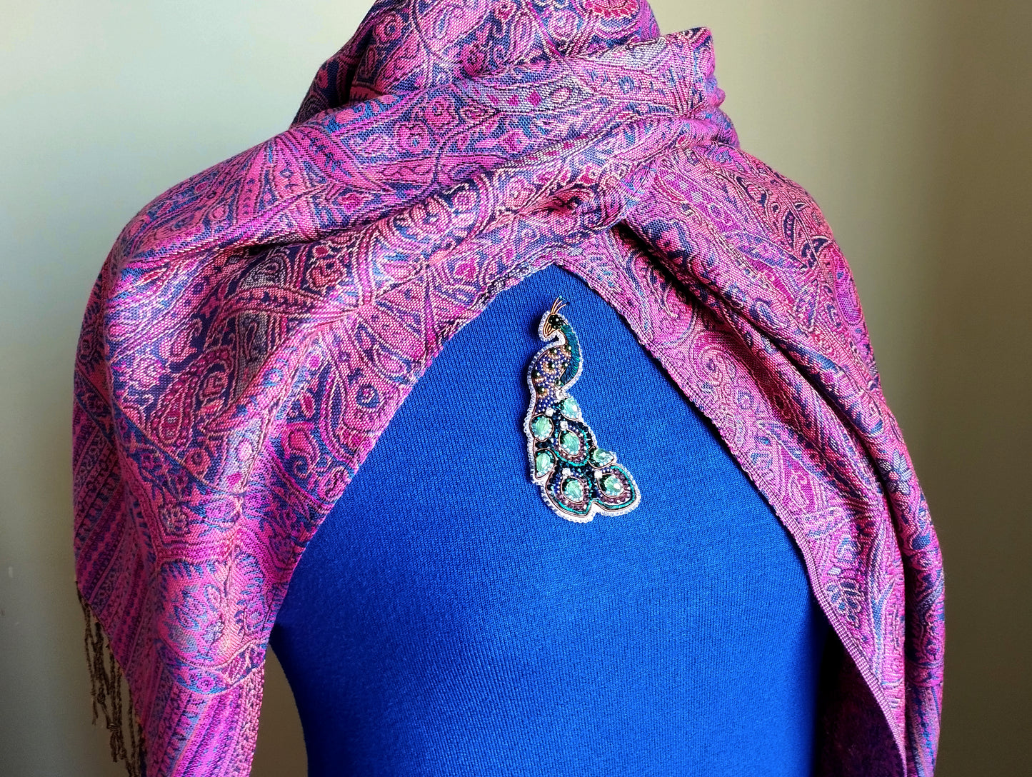 A mannequin wearing a blue top with a pink and purple paisley patterned shawl draped over it, and a decorative brooch on the chest.
