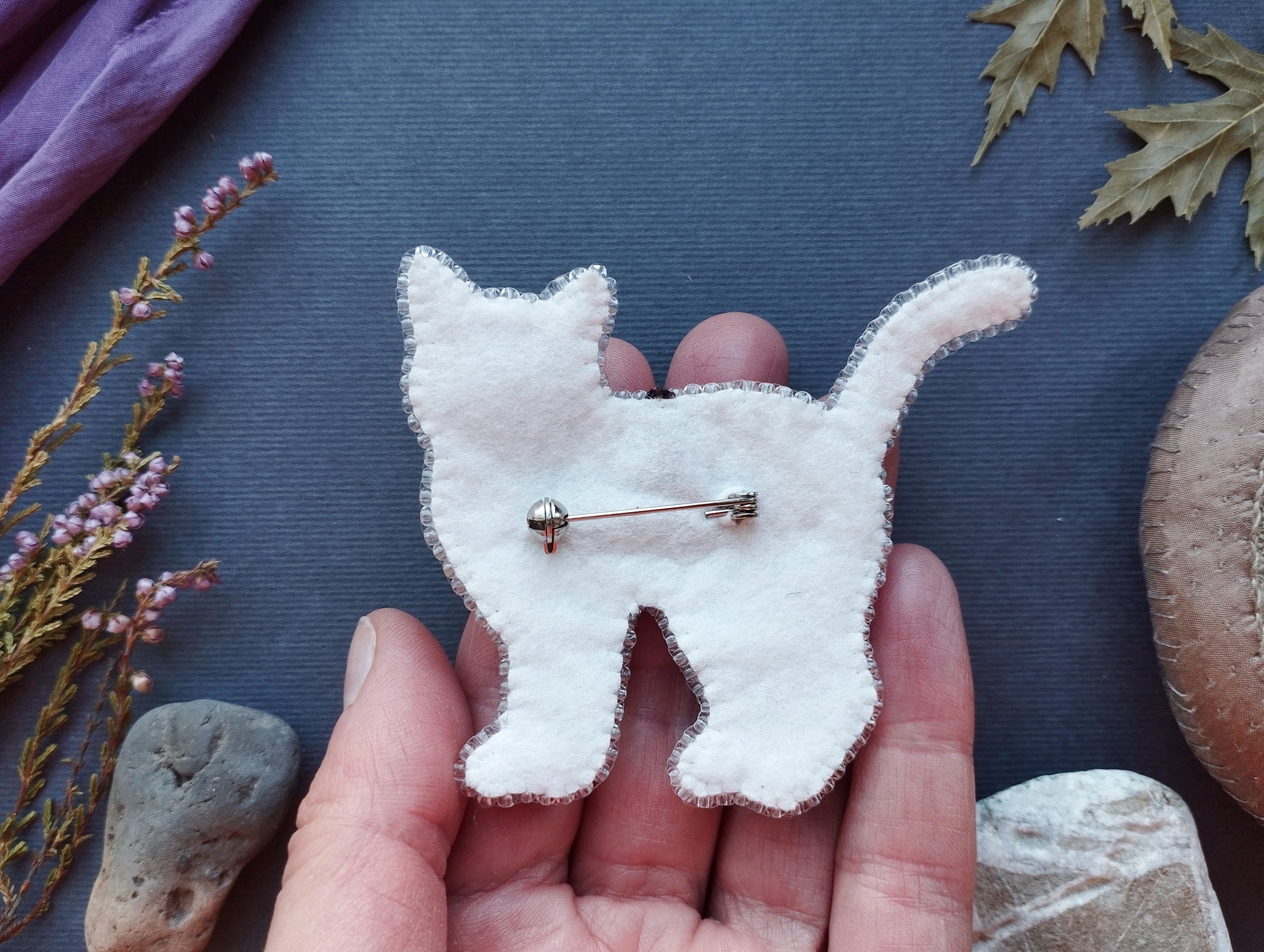 A hand holding a white felt cat pin against a dark background with various natural elements, such as leaves and rocks.