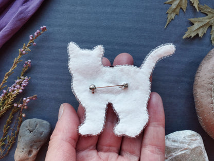 A hand holding a white felt cat pin against a dark background with various natural elements, such as leaves and rocks.