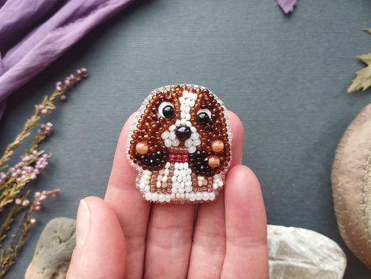 A hand holding a small, intricately beaded embroidered dog-shaped brooch against a gray background.