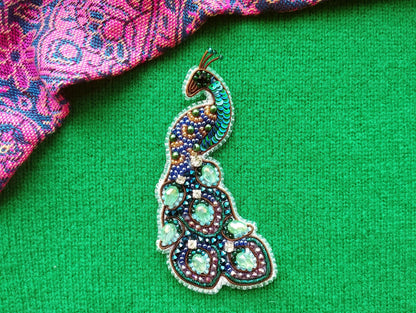 A colorful, intricately designed peacock brooch is displayed against a vibrant green background.