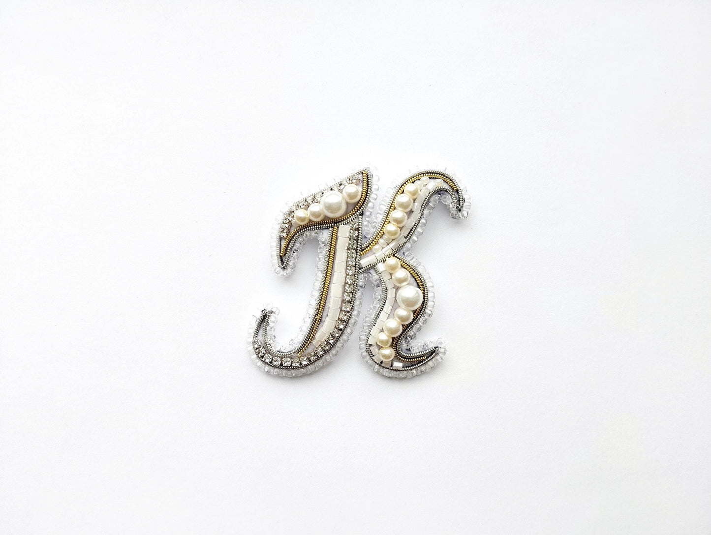 A decorative letter 'K' made of beads, pearls, and metallic elements, placed against a white background.