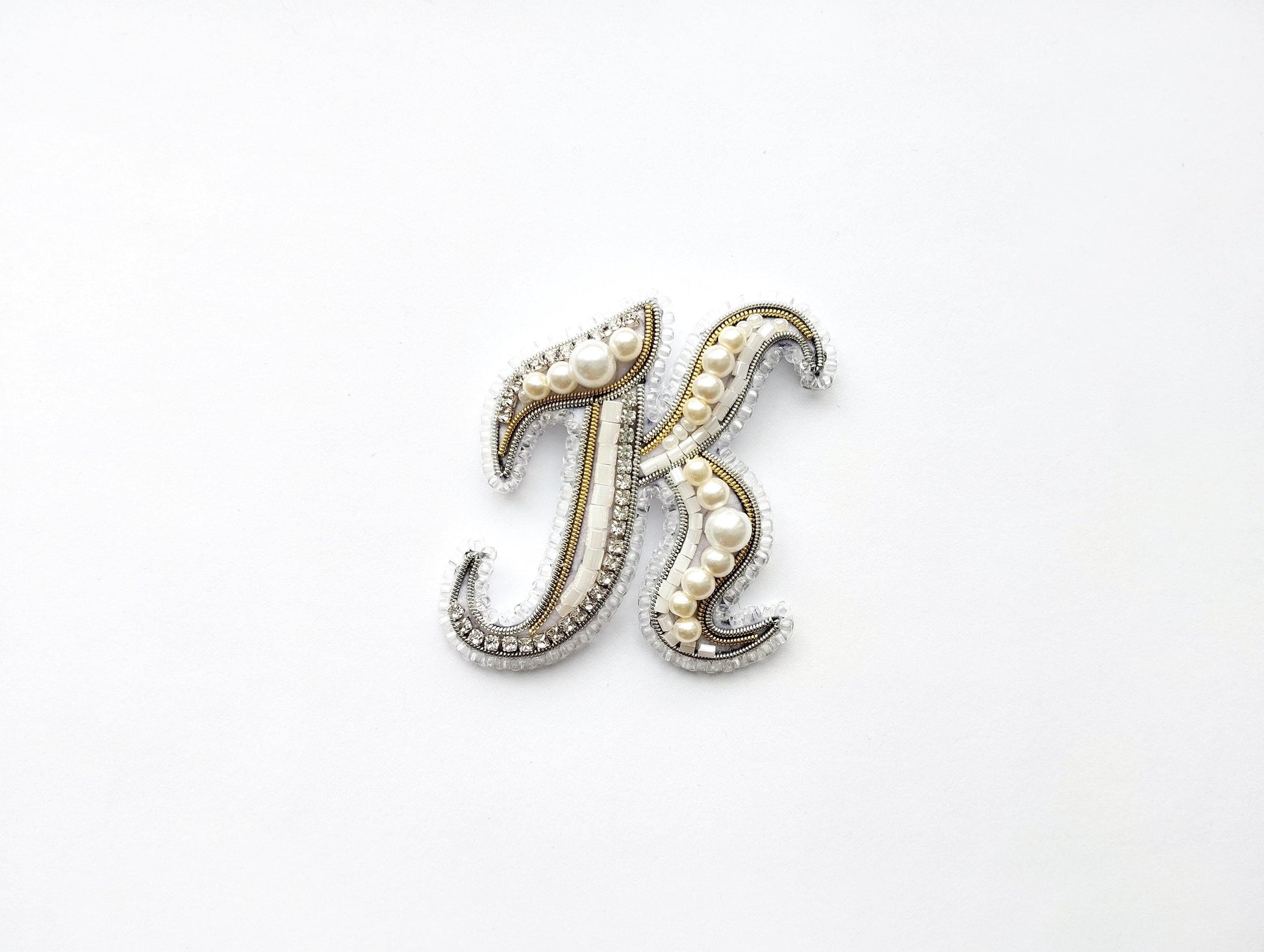 A decorative letter 'K' made of beads, pearls, and metallic elements, placed against a white background.