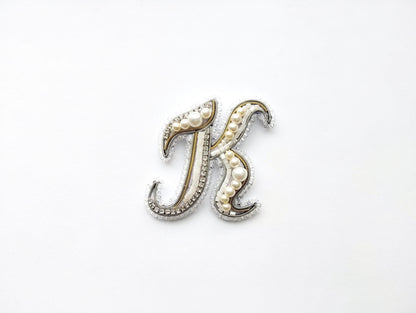 A decorative letter 'K' made of beads, pearls, and metallic elements, placed against a white background.