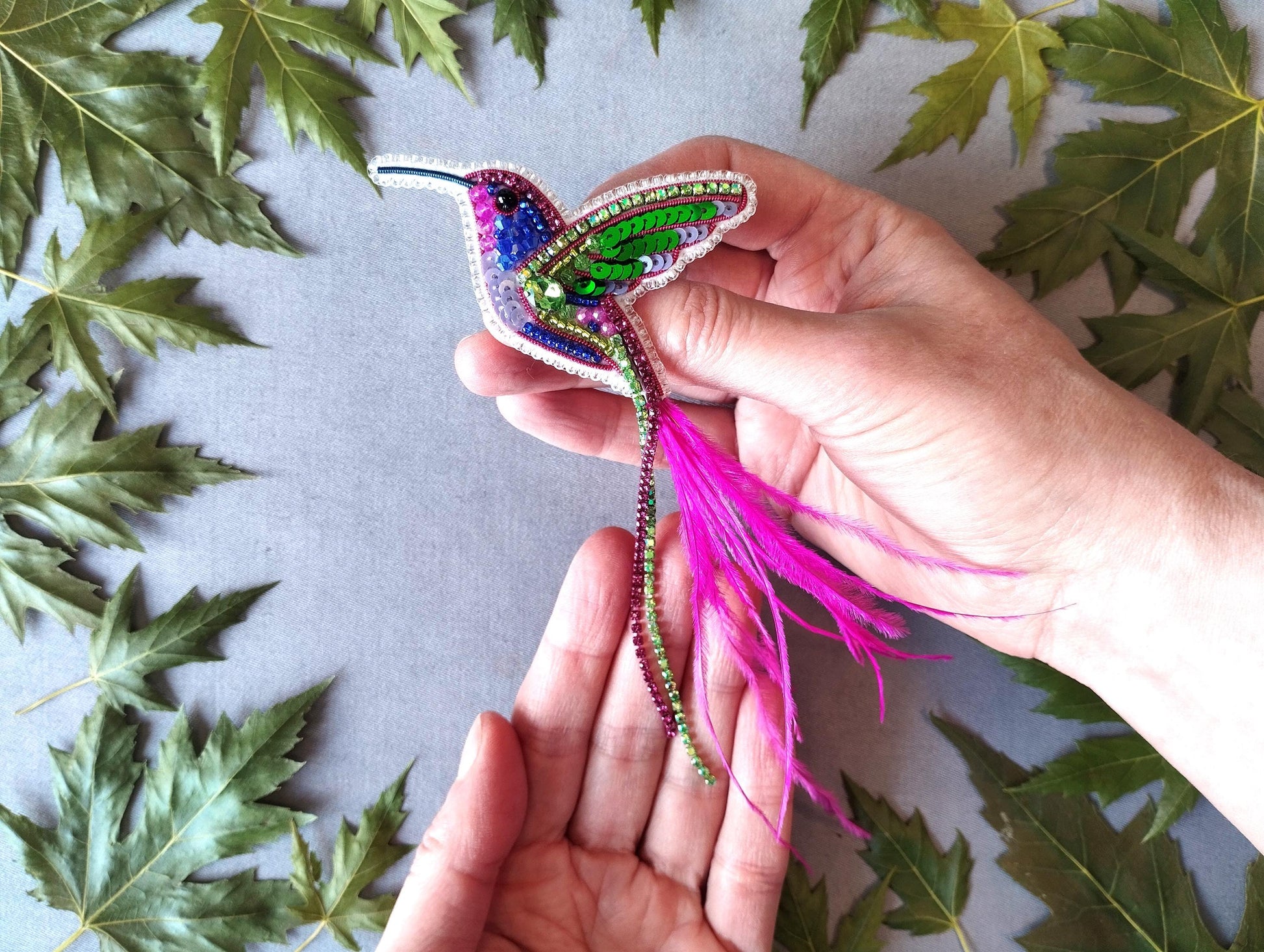 A pair of hands holding a colorful, beaded hummingbird brooch against a backdrop of green leaves.
