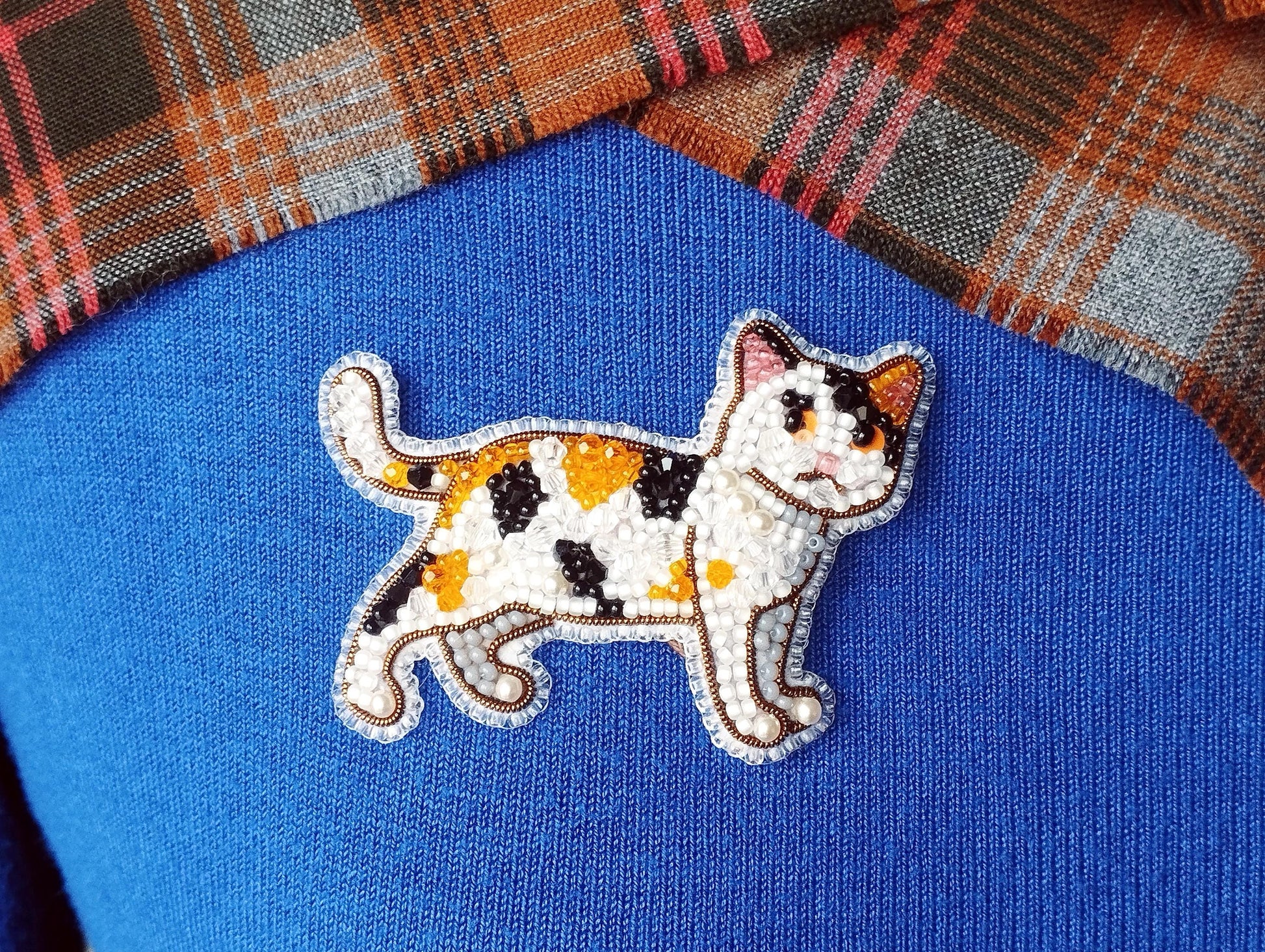 A close-up image of a cat embroidered on a blue fabric, with a plaid fabric visible in the background.