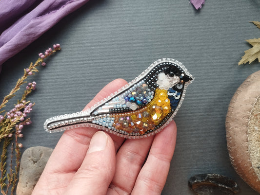 A hand is holding a decorative bird brooch made of colorful beads and sequins, with a purple fabric and some dried flowers in the background.