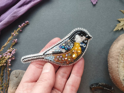 A hand is holding a decorative bird brooch made of colorful beads and sequins, with a purple fabric and some dried flowers in the background.