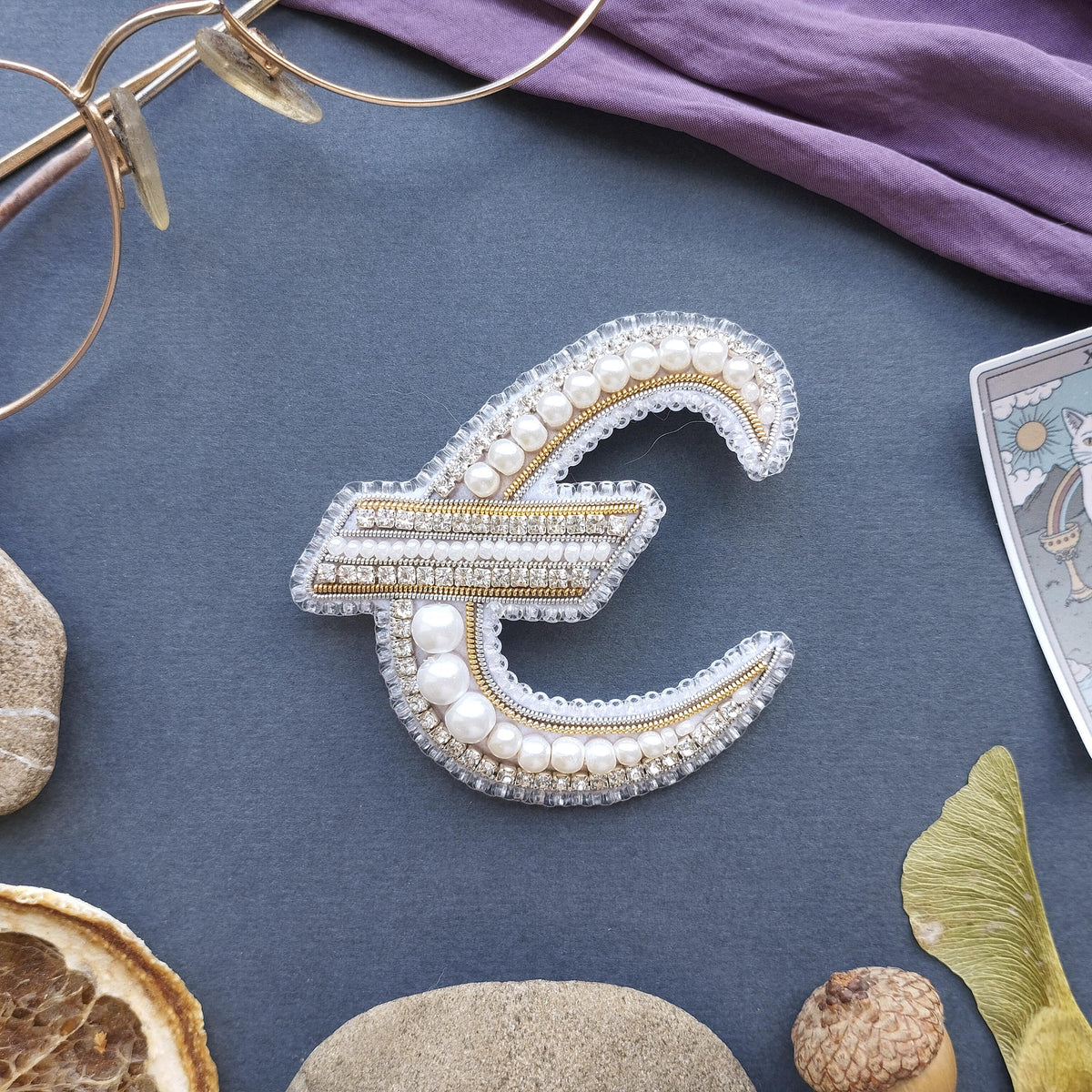 a close-up view of a decorative letter 'E' made of pearls and diamonds, surrounded by various objects such as a pair of glasses, a purple fabric, and some natural elements like a leaf and a rock.