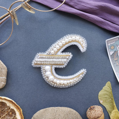 a close-up view of a decorative letter 'E' made of pearls and diamonds, surrounded by various objects such as a pair of glasses, a purple fabric, and some natural elements like a leaf and a rock.