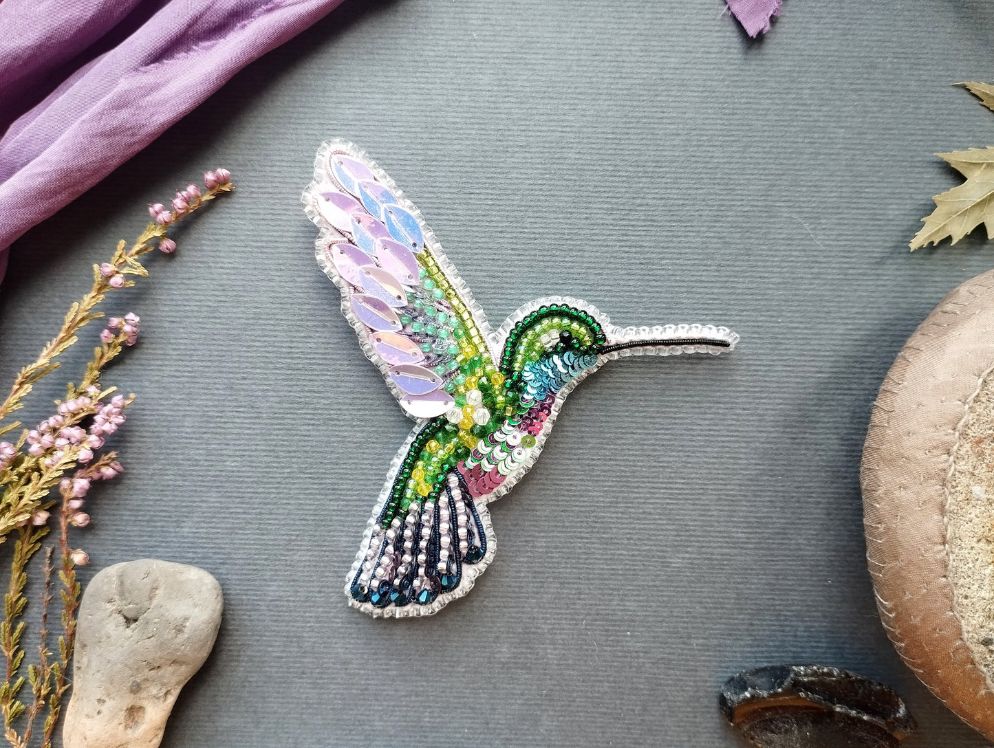a colorful, intricately designed hummingbird brooch or pin, surrounded by various natural elements such as flowers, leaves, and rocks.