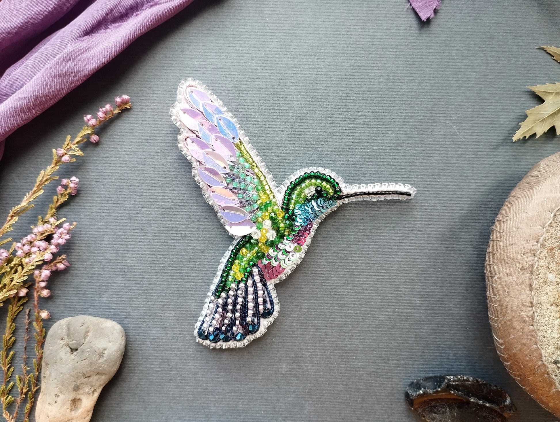 a colorful, intricately designed hummingbird brooch or pin, surrounded by various natural elements such as flowers, leaves, and rocks.