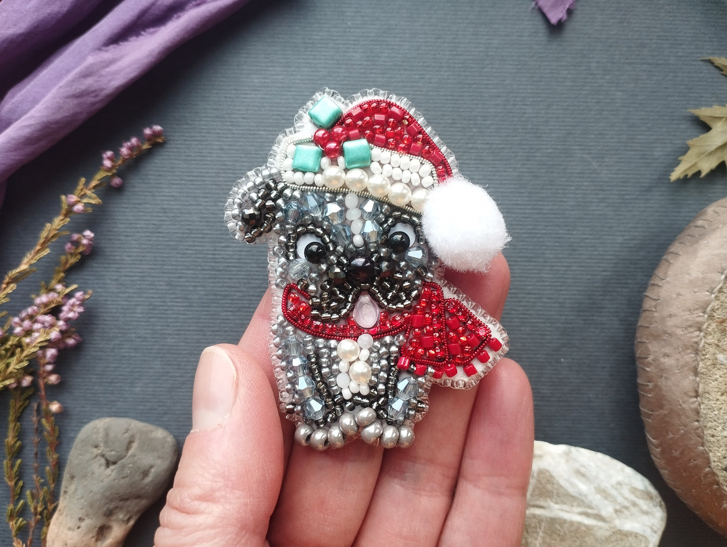 A hand holding a small, intricately beaded embroidered dog wearing a Santa hat and scarf.