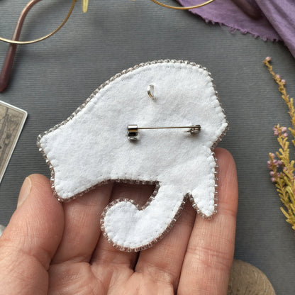 A hand holding a white felt object with a silver pin attached to it, placed on a gray surface with some flowers and a pair of glasses in the background.