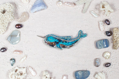 a blue and silver brooch with a whale design, surrounded by various natural elements such as pearls, seashells, and crystals.