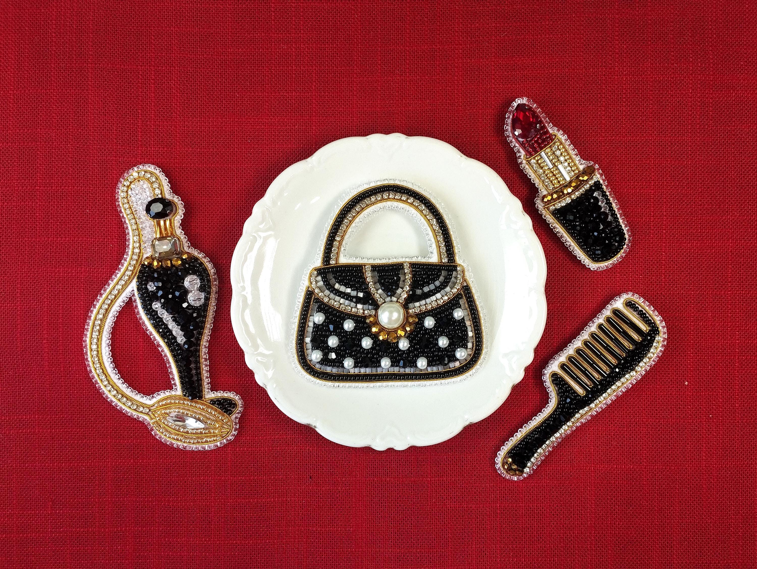 a white plate with a black and gold handbag, a red lipstick, and a black comb, all of which are intricately designed and embellished with jewels and pearls.