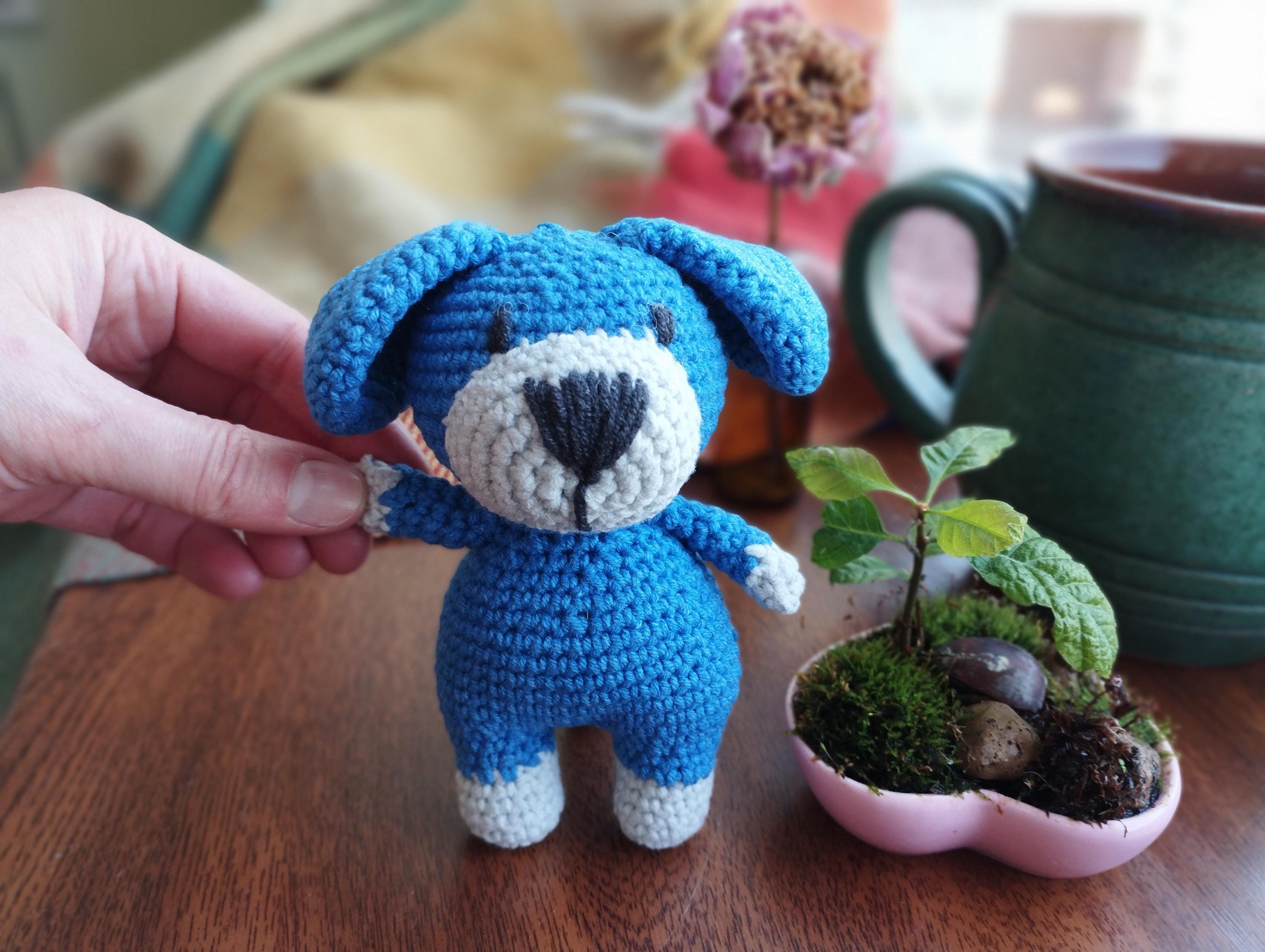 a crocheted blue teddy bear next to a small plant